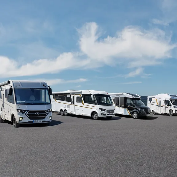 Reihe von sechs Wohnmobilen verschiedener Modelle und Größen, geparkt auf einem großen Asphaltplatz am Meer bei klarem Himmel.