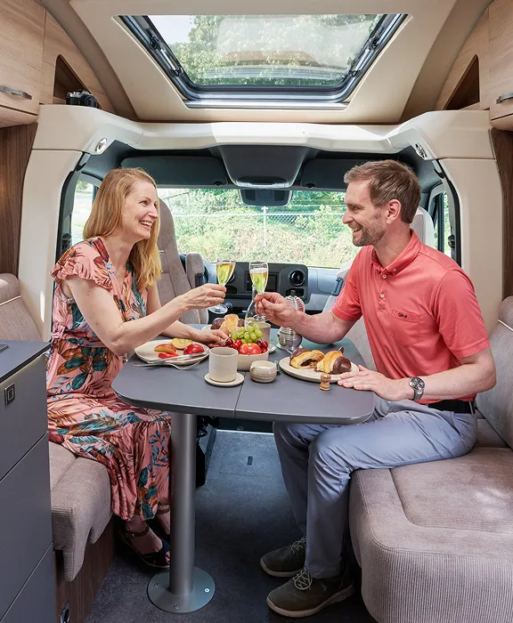 Paar genießt ein gemeinsames Frühstück mit Sekt im gemütlichen Innenraum eines modernen Wohnmobils mit Küchenzeile und großer Fensterfront.