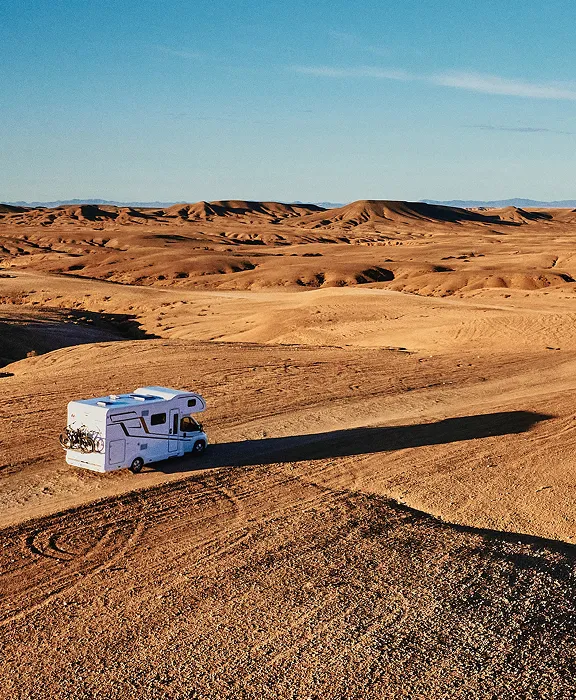 Wohnmobil fährt auf einer einsamen Straße durch eine weite, hügelige Wüstenlandschaft unter klarem blauem Himmel.